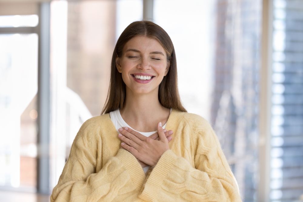 Grateful Woman Smiling Warmly – Orthodontist in Arcadia CA A young woman stands indoors with her hands over her chest, smiling gently with straight, healthy teeth that reflect confidence and comfort – Orthodontist in Arcadia CA.