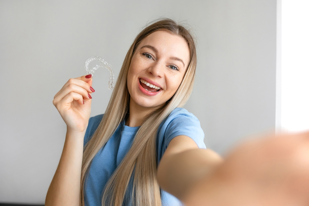 A cheerful young woman shows off her clear Invisalign aligner while smiling brightly, celebrating her progress toward a straighter smile – Arcadia Invisalign