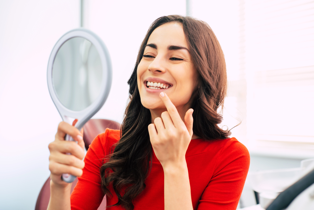 A joyful woman looks into a handheld mirror and points at her straight teeth, celebrating her Invisalign treatment progress – Invisalign in San Gabriel.