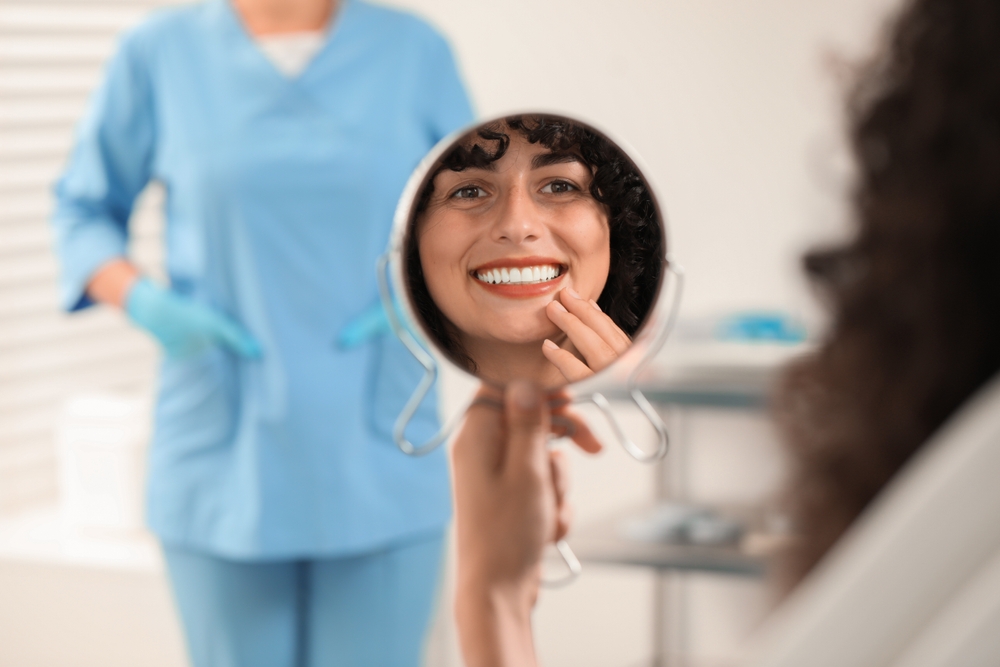 A patient happily examines her bright, straight smile in a hand mirror at the dental office, reflecting the confidence that comes with Invisalign treatment – Arcadia Invisalign.