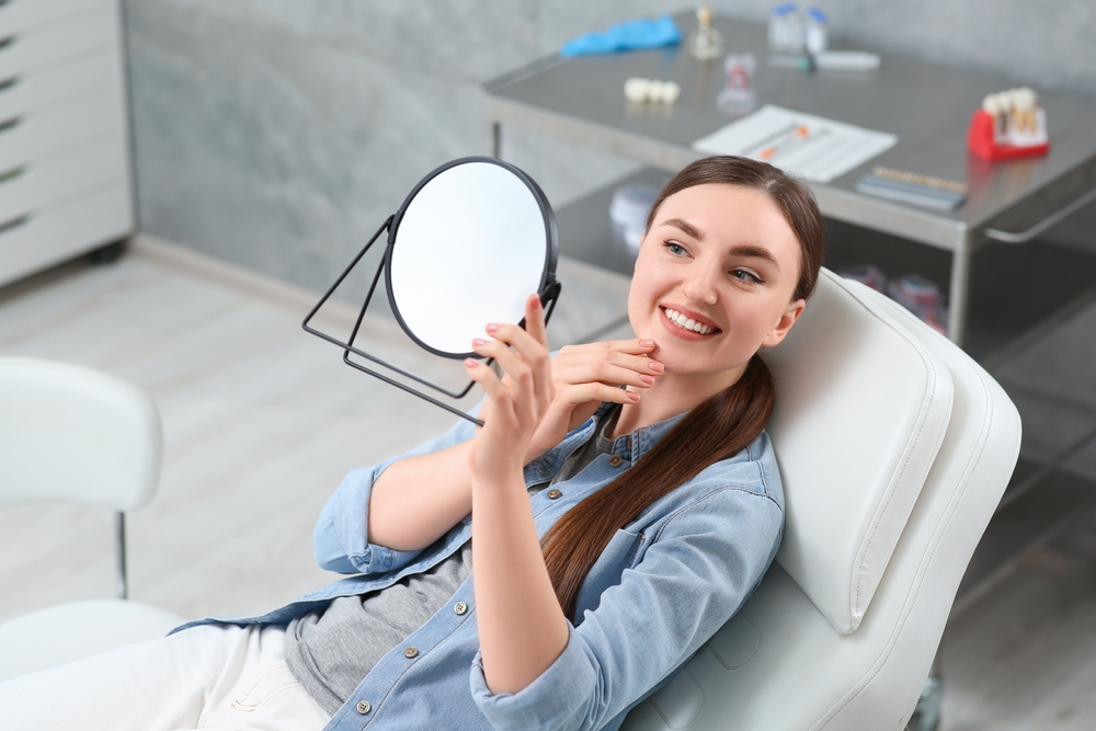 A smiling woman relaxes in the dental chair while examining her teeth in a handheld mirror, appreciating her Invisalign results – Invisalign Alhambra.