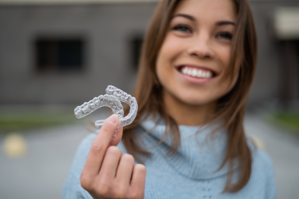 A smiling woman holds up her clear aligners outside, proudly showcasing the convenience and discreet design of Invisalign – Invisalign in Pasadena